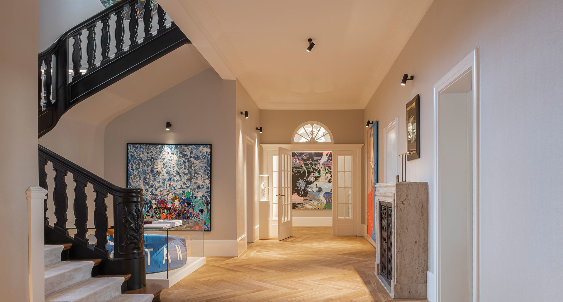 Elegant hallway with herringbone parquet, black staircase, artwork on walls, and Artemide adjustable ceiling spotlights illuminating the space