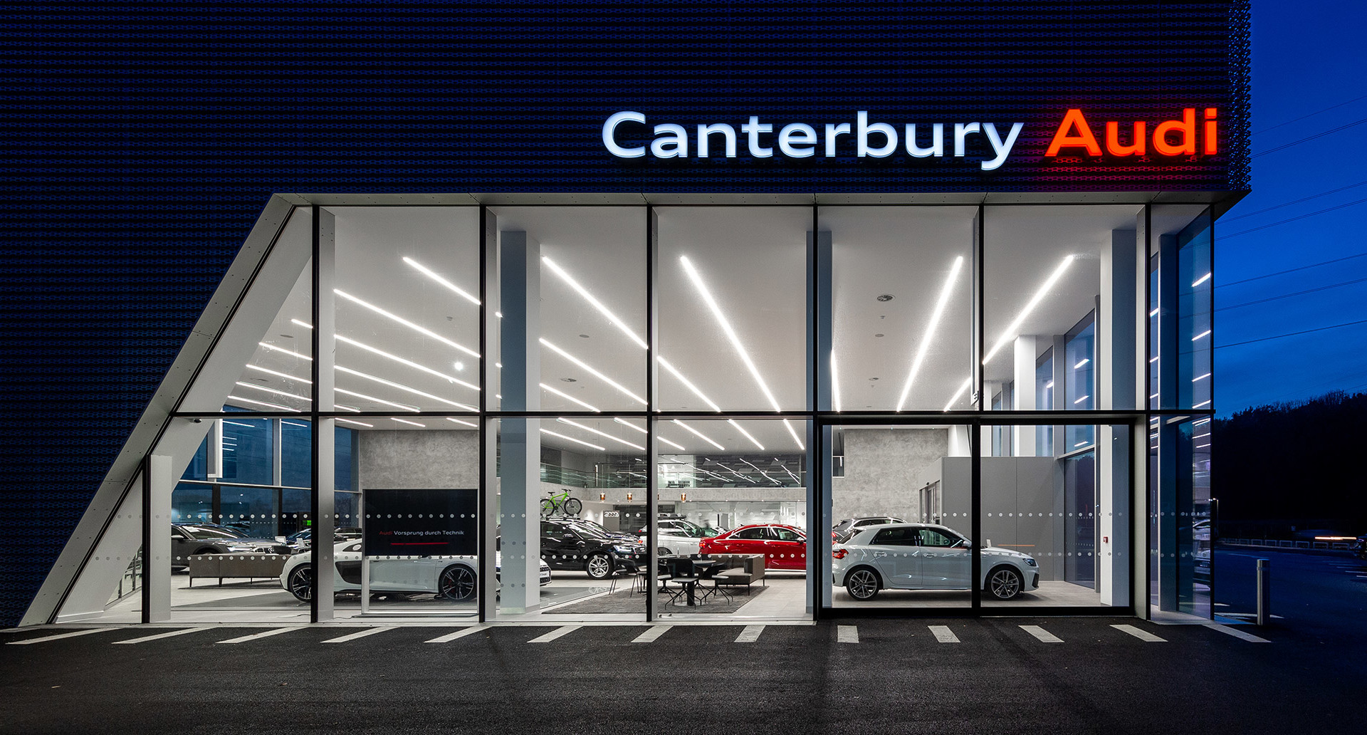 Showroom Canterbury Audi di notte con luci a soffitto LED lineari che illuminano veicoli di lusso dietro una facciata in vetro dal pavimento al soffitto