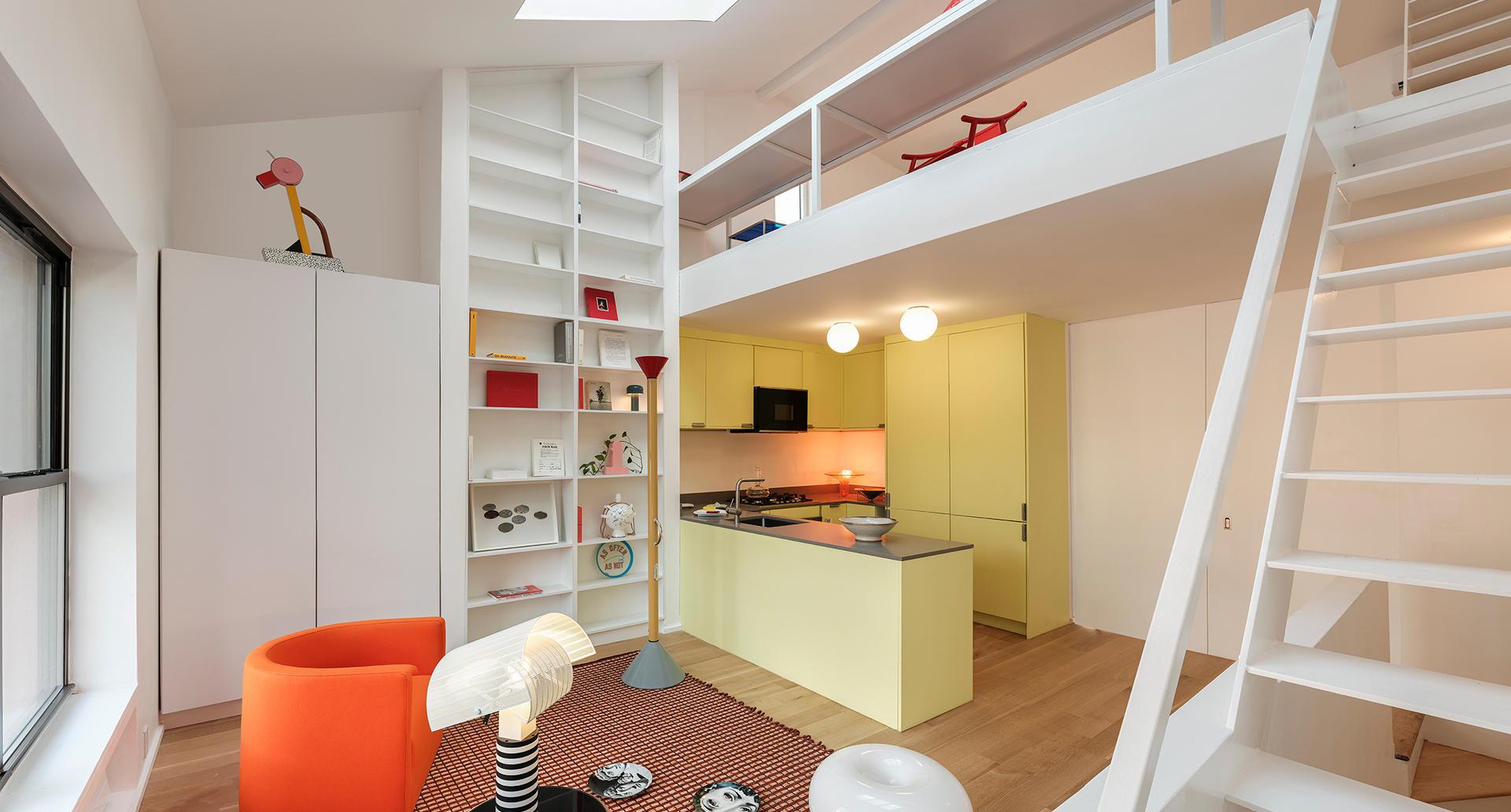 Interno di loft moderno con lampade Artemide Snoopy e Tolomeo, cucina gialla, soppalco, scaffalatura bianca e poltrona arancione.