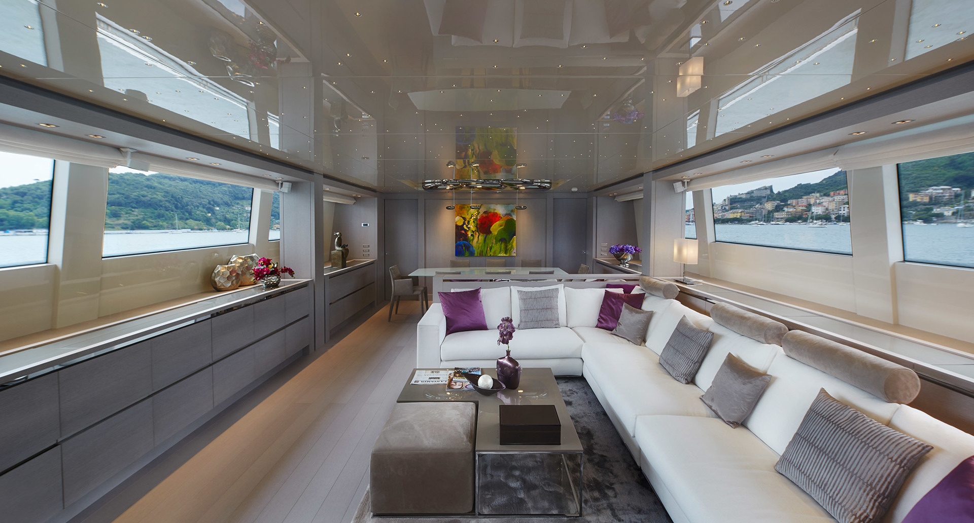 Salone di yacht di lusso con divano componibile bianco, accenti viola, finestre panoramiche e elegante illuminazione a sospensione sopra il tavolino.