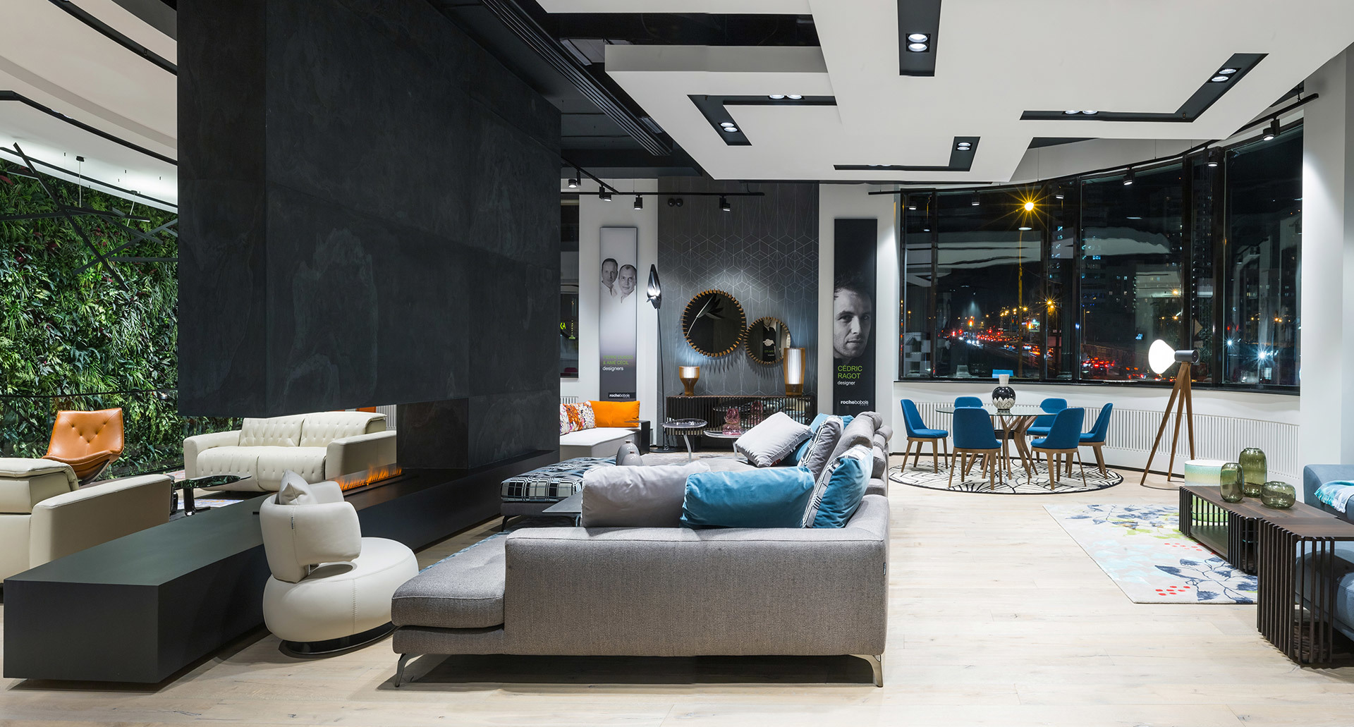 Showroom moderno con faretti da soffitto, lampada da terra su treppiede, divano grigio, camino, parete verde e vista città di notte