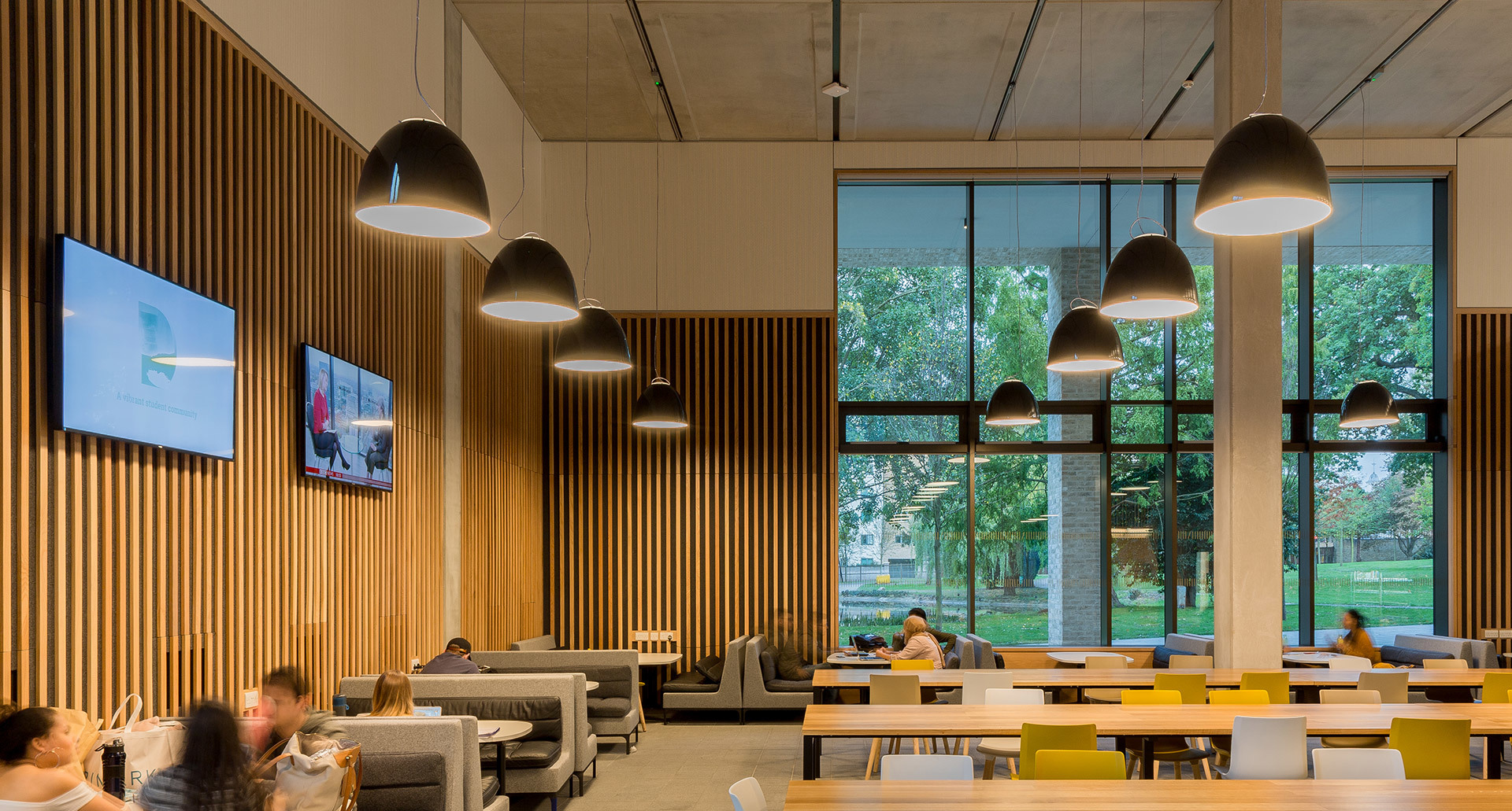 Modern cafeteria with black dome pendant lights over dining tables, wood-slat walls, large windows, and colorful seating areas.
