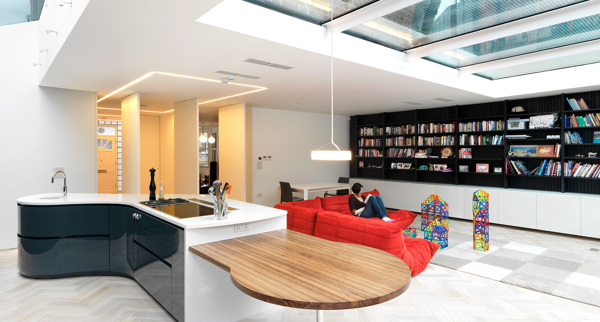 Soggiorno open space moderno con lampada a sospensione Artemide sopra l'isola cucina, soffitto con lucernario, divano rosso e libreria a tutta altezza.