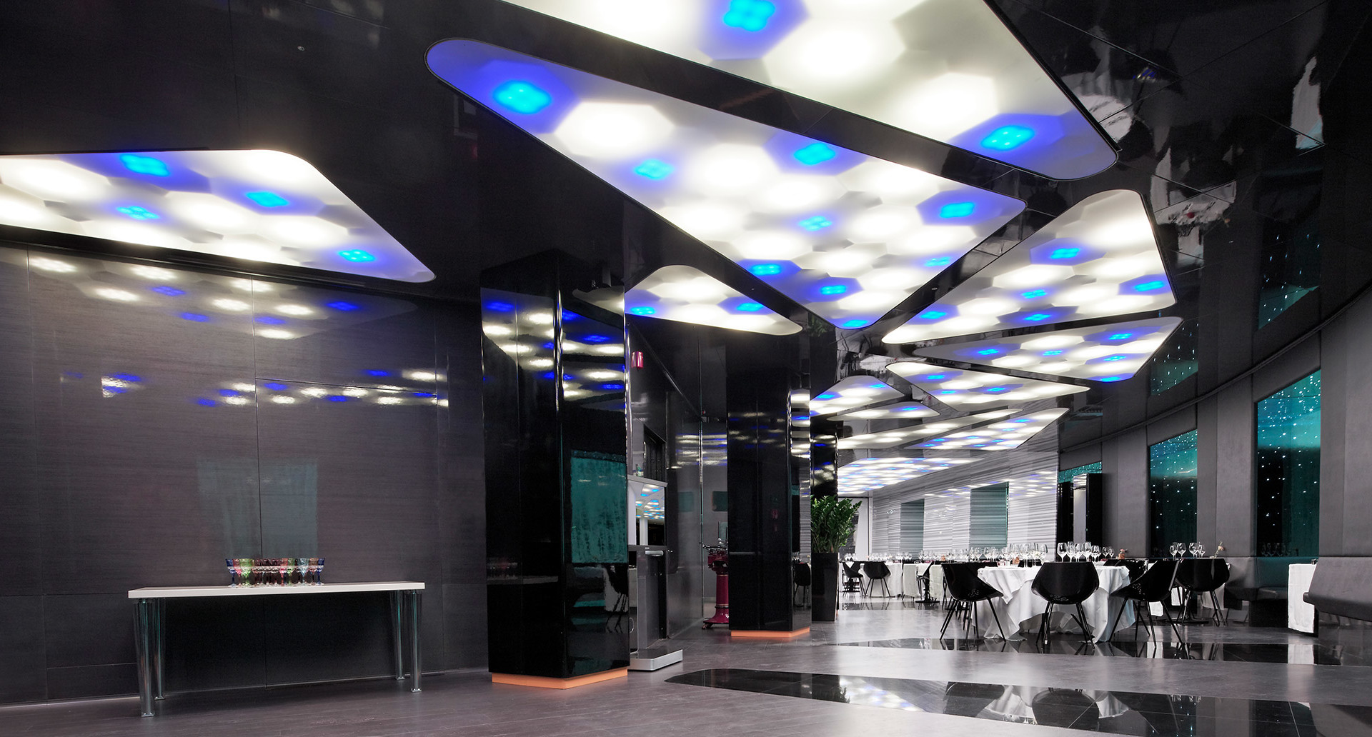 Ingresso moderno con pannelli soffitto geometrici a LED bianchi e blu, riflessi su pavimenti scuri lucidati, con area ristoro sullo sfondo
