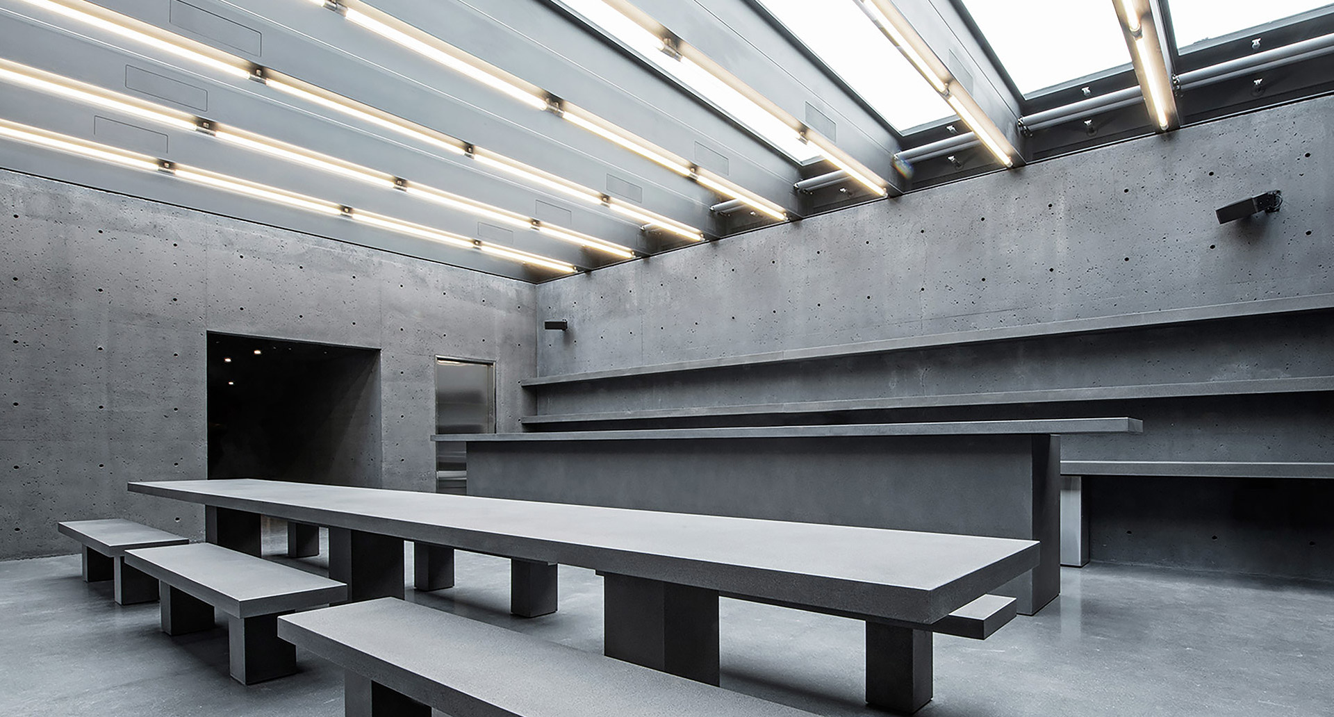 Minimalist concrete hall with long gray tables, benches, and Artemide linear LED lights integrated into a skylight ceiling structure.