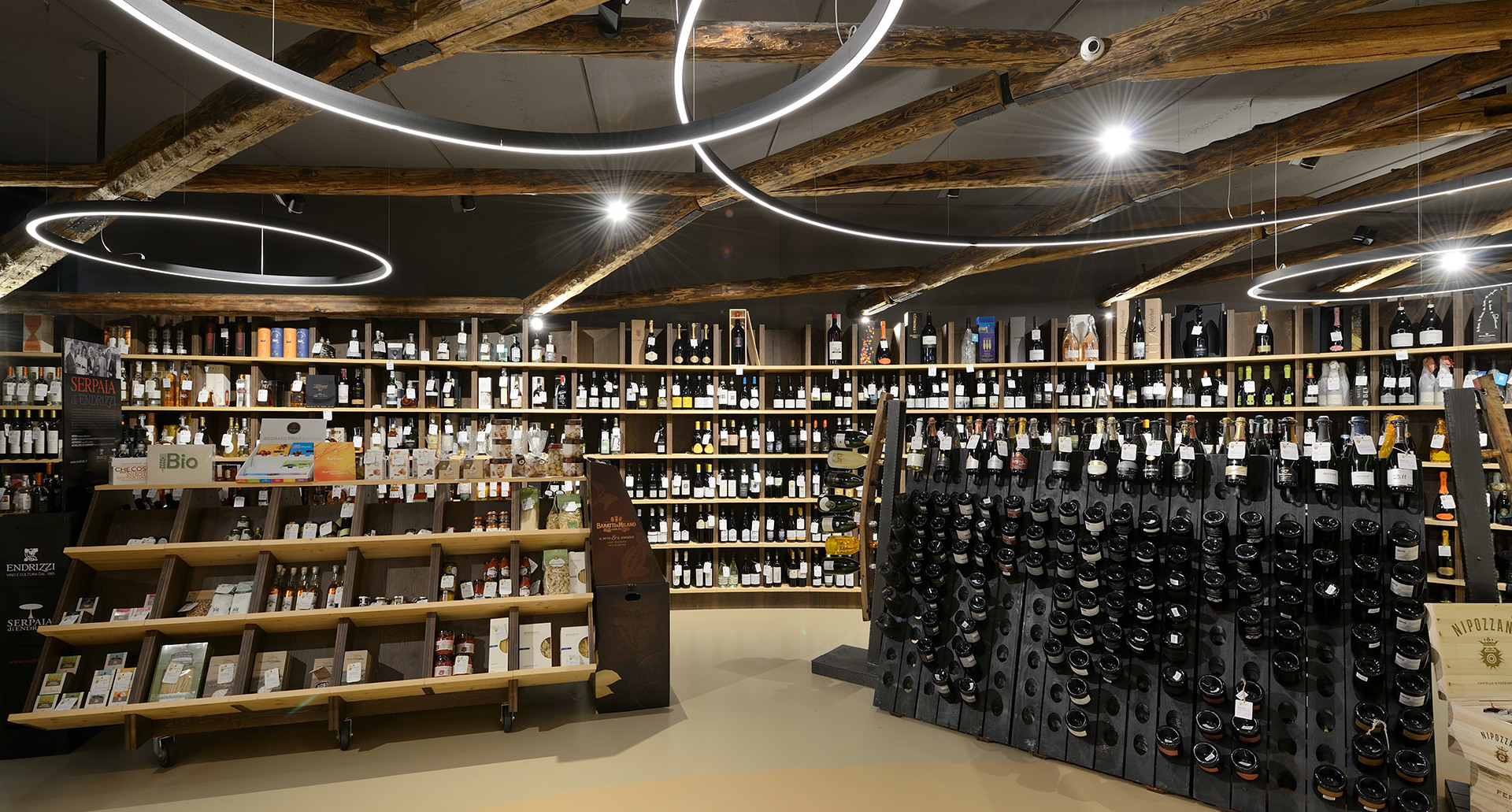 Interno di enoteca con lampade a sospensione ad anello LED circolari che illuminano scaffali in legno con bottiglie di vino.