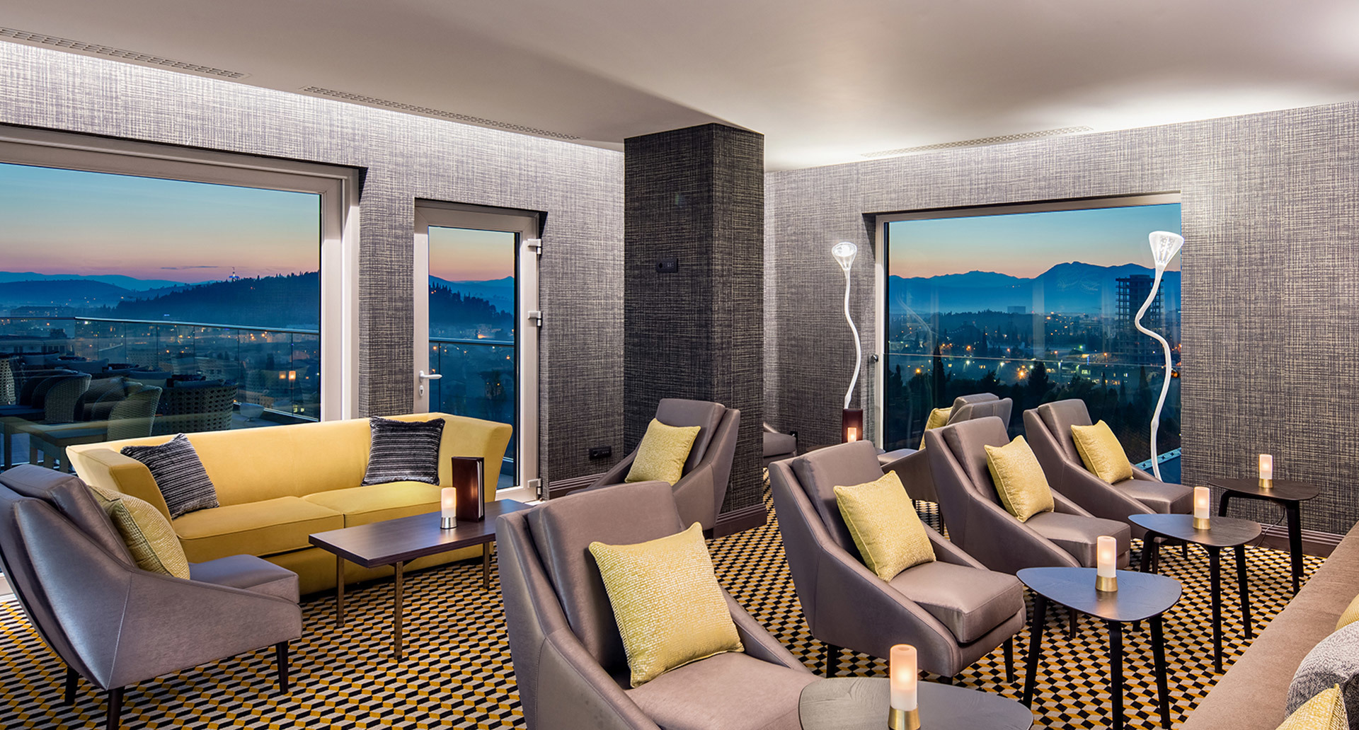 Elegante lounge di hotel al crepuscolo con lampade da terra Artemide, divani gialli, poltrone grigie, tappeto fantasia e vista panoramica sulla città