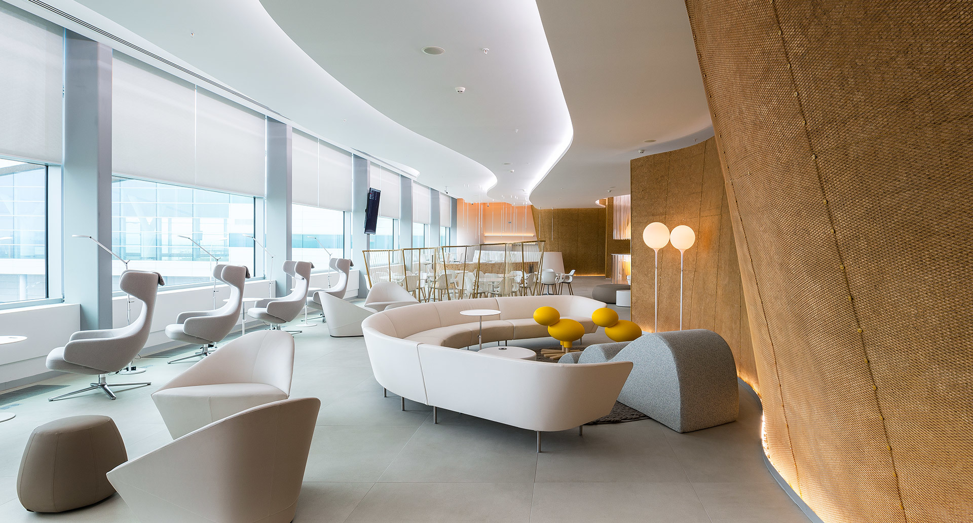 Lounge moderna con divani bianchi curvi, accenti gialli, lampade da terra Artemide e illuminazione indiretta a soffitto che crea un'atmosfera calda.