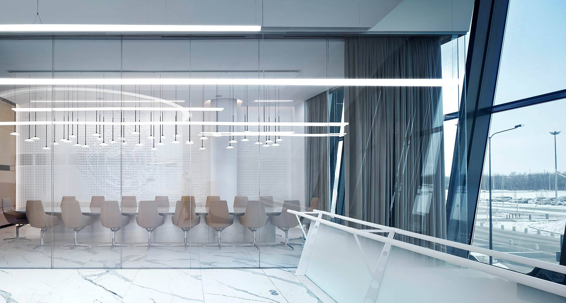 Sala conferenze moderna con pavimenti in marmo, pareti in vetro e luci a sospensione lineari che illuminano un lungo tavolo con sedie beige.