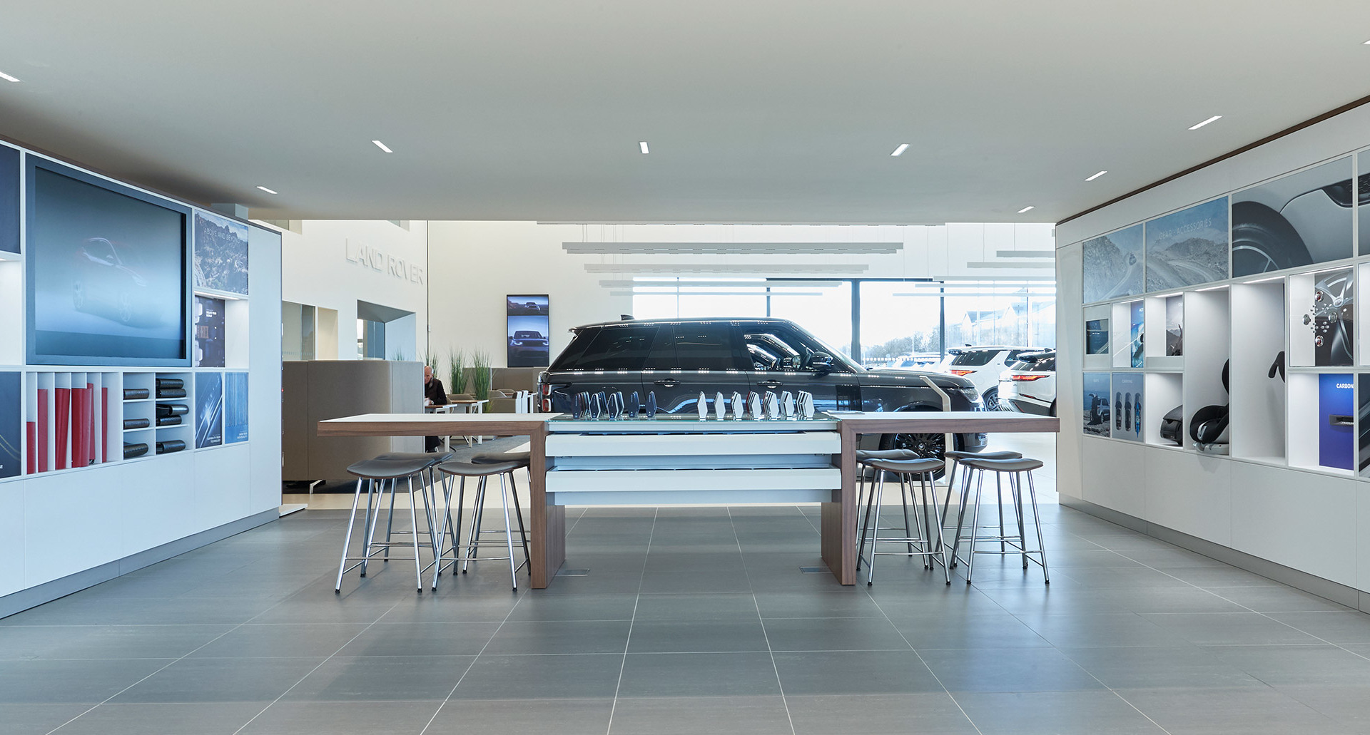 Modern car showroom with recessed ceiling lights, a central display table, bar stools, and a dark SUV on display.