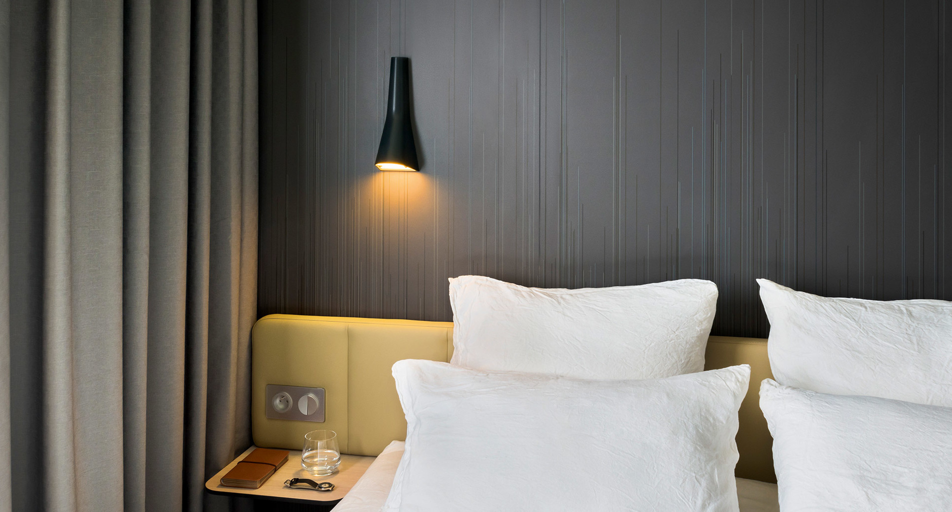 Lampada da parete conica nera che proietta luce calda verso il basso sopra la testiera di un letto d'albergo con cuscini bianchi e pannello in legno grigio.