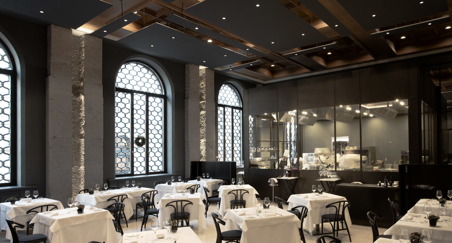 Elegant restaurant interior with Artemide recessed spotlights illuminating white-clothed tables, dark walls, arched windows, and exposed wooden ceiling beams.