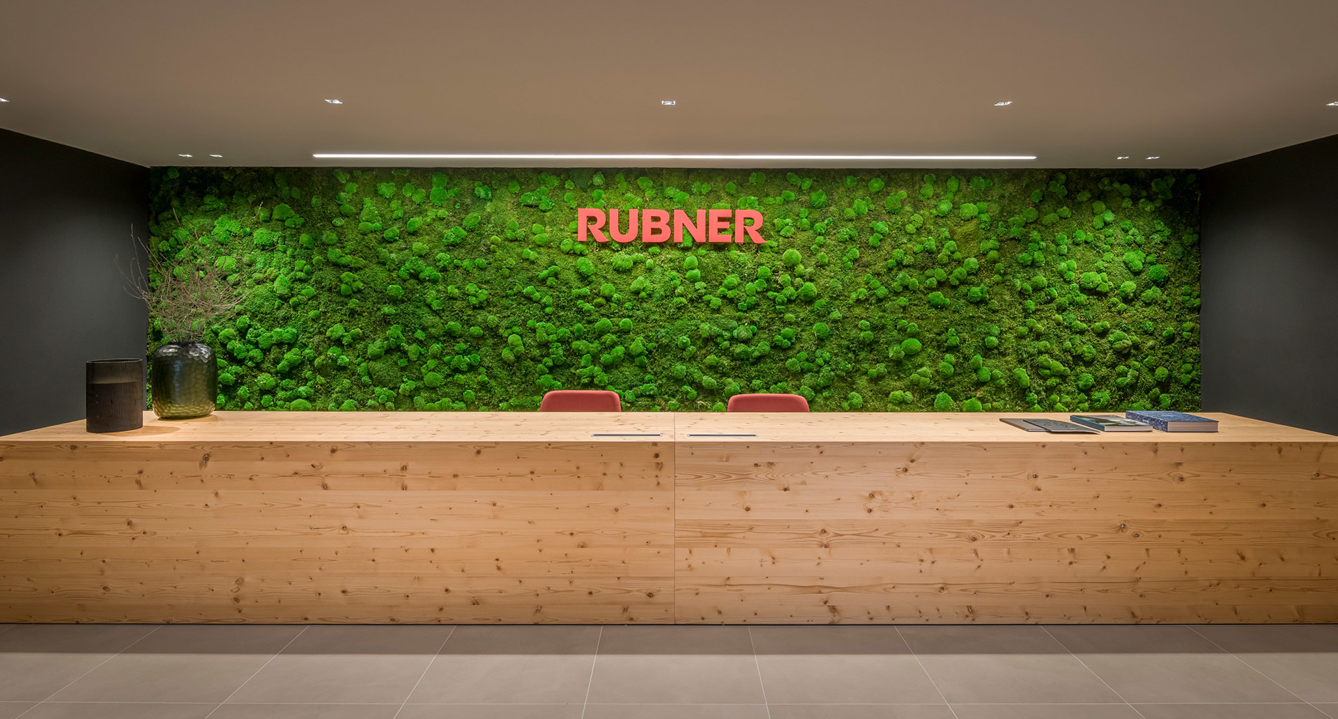 Reception Rubner con bancone in legno naturale e parete di muschio verde, illuminata da faretti a soffitto incassati.
