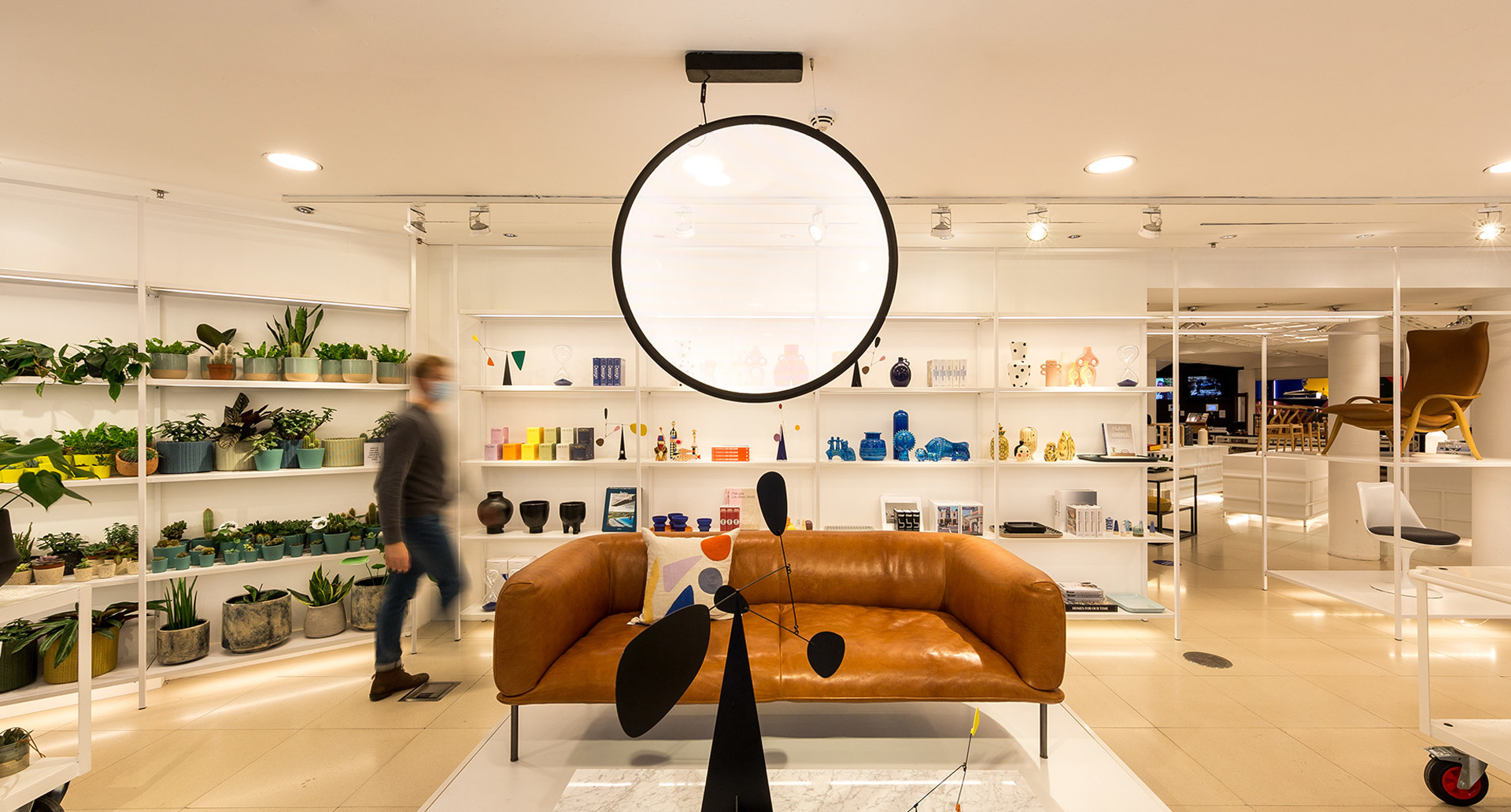 Lampada a sospensione circolare Artemide illumina uno showroom moderno con divano in pelle, scaffali colorati, piante e una scultura mobile.