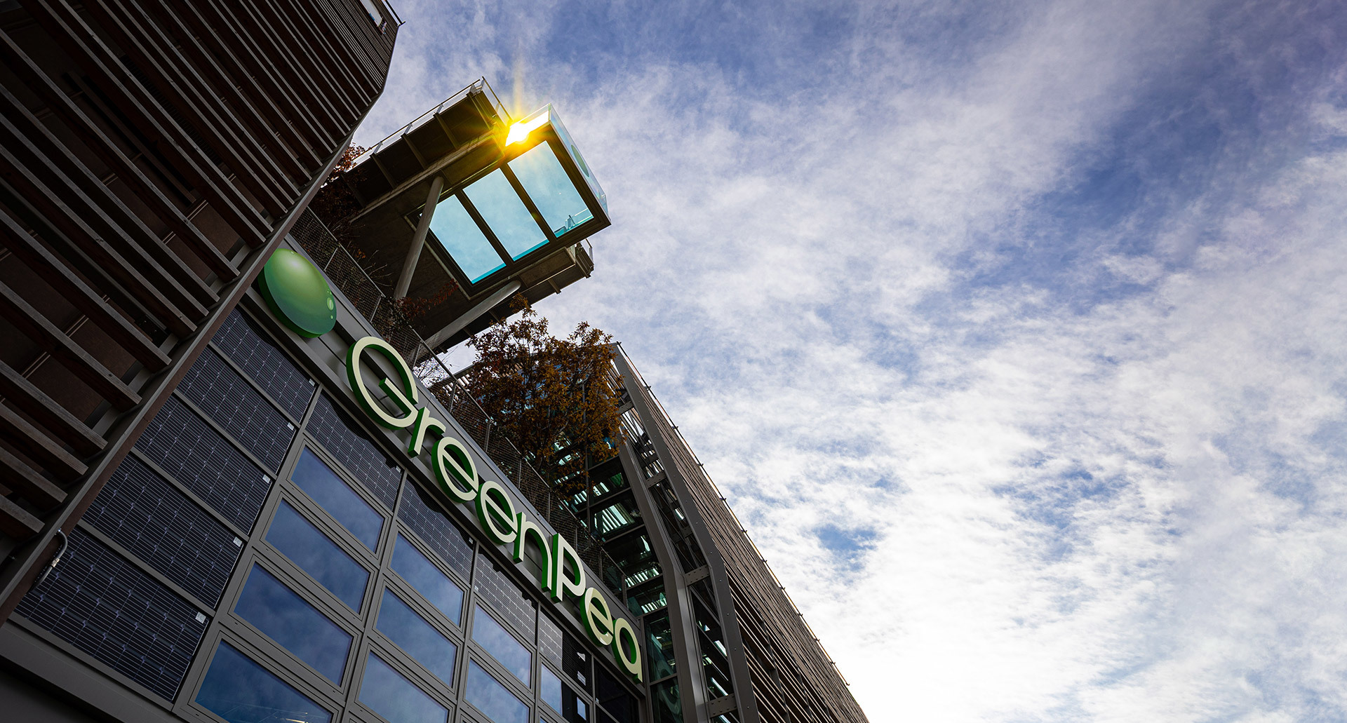 Low-angle view of GreenPea building facade with solar panels, rooftop garden, and a bright outdoor light fixture against a cloudy sky.