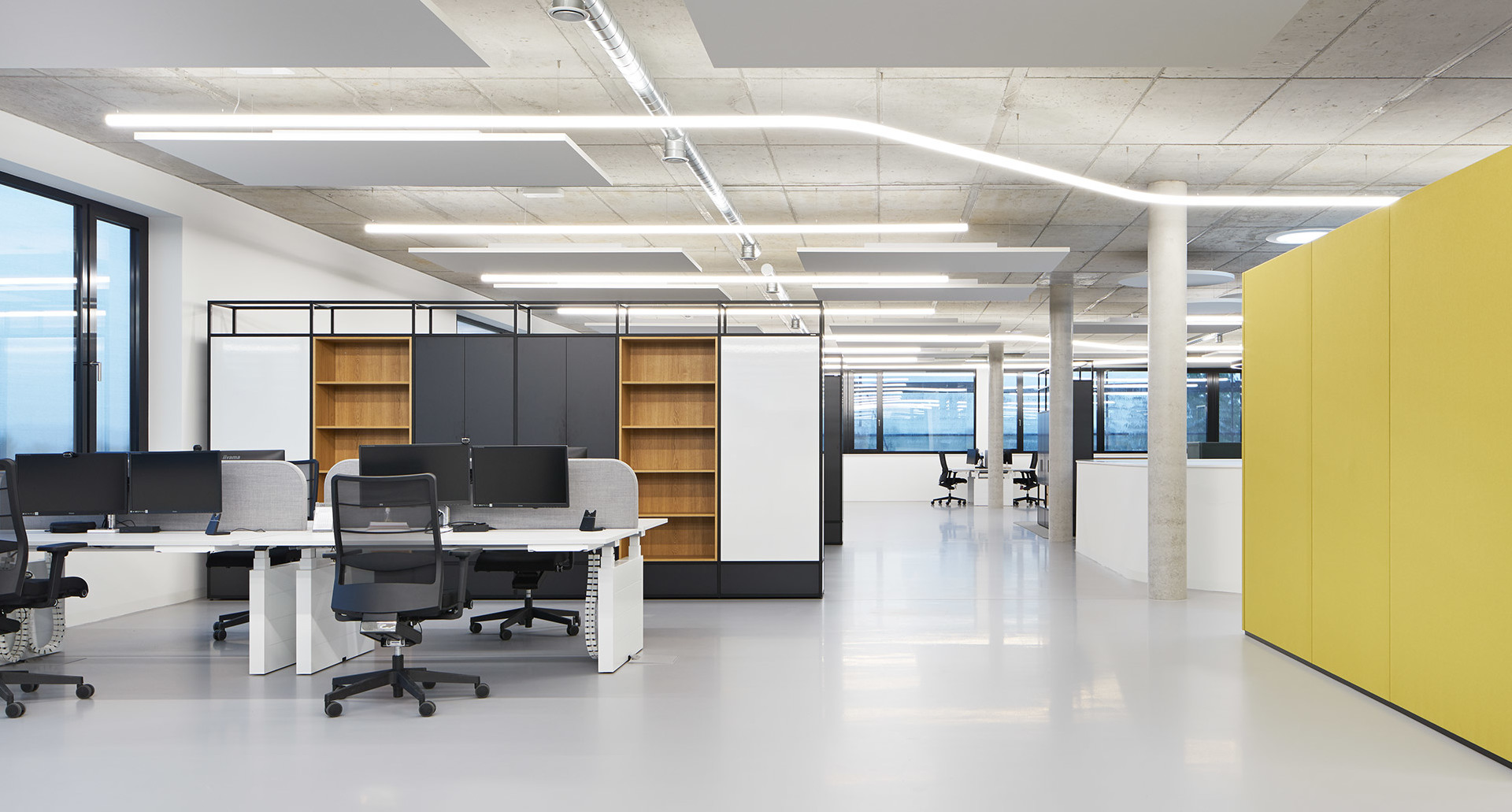 Ufficio open space moderno con lampade lineari LED a soffitto Artemide che illuminano postazioni di lavoro, librerie e pannelli divisori gialli.