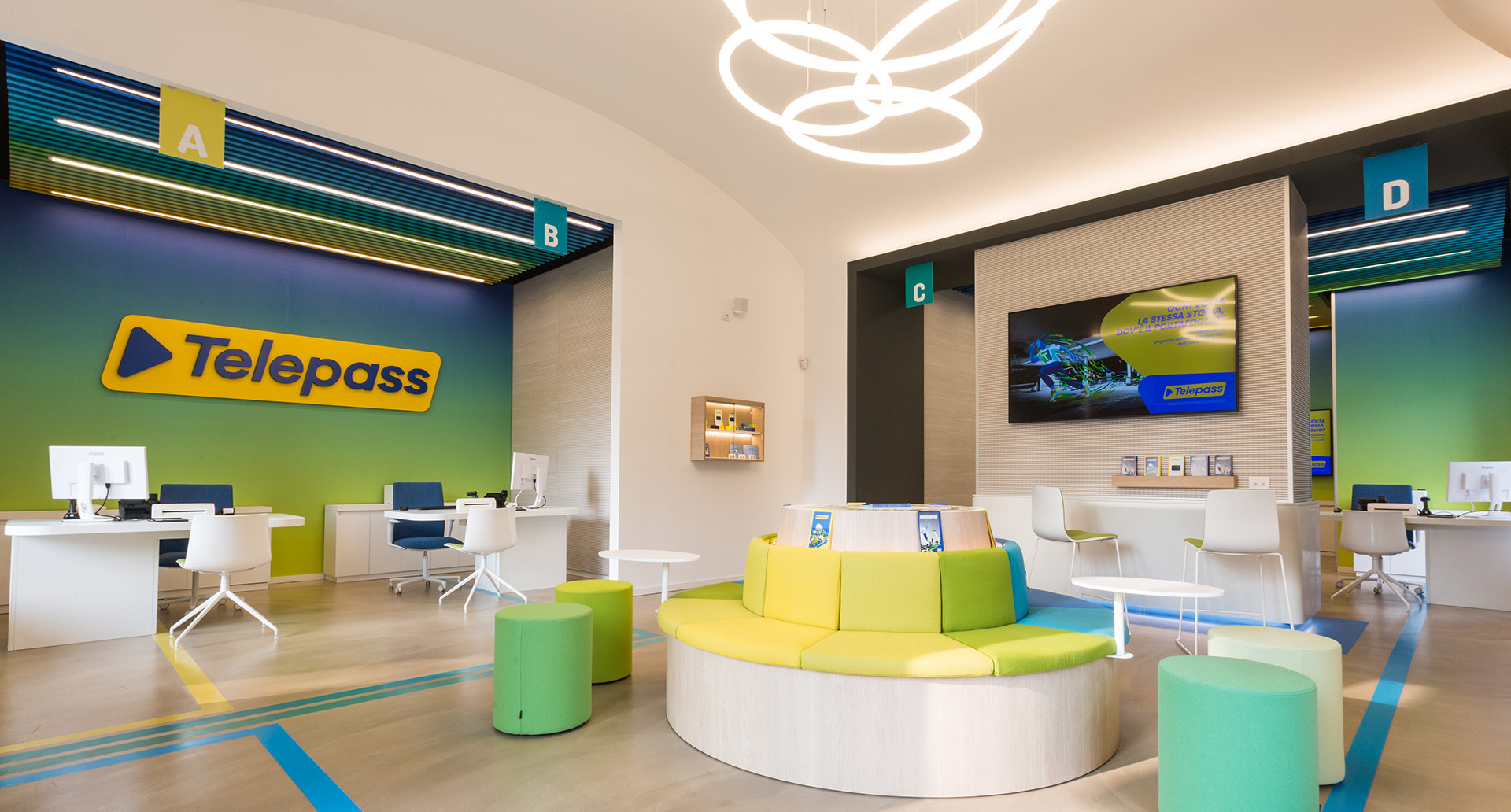 Interno del negozio Telepass con lampade a sospensione circolari Artemide che illuminano uno spazio moderno e colorato con sedute verdi e gialle.