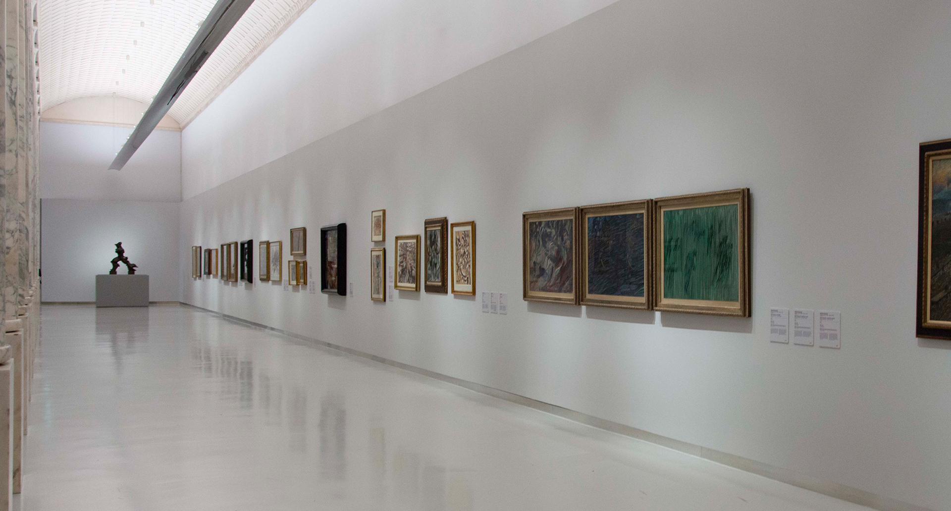 Corridoio di galleria d'arte con illuminazione a soffitto incassata che illumina dipinti su pareti bianche e una scultura in fondo.