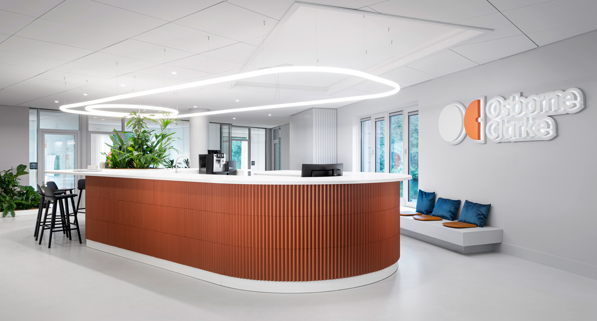 Reception uffici Osborne Clarke con lampada a sospensione ad anello ovale Artemide che illumina una scrivania curva con pannelli in legno e piante verdi.