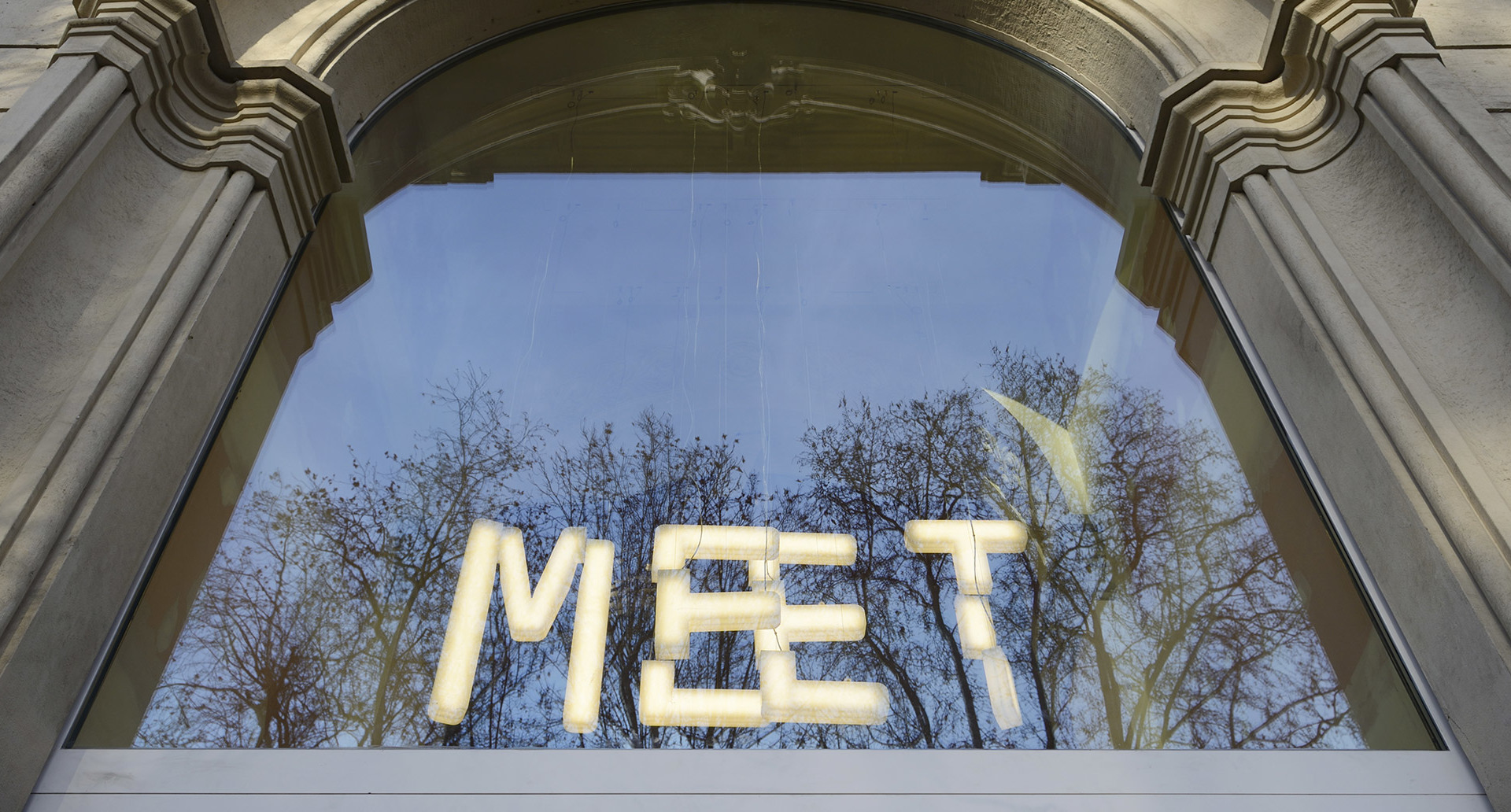 Illuminated 'MEET' sign made of glowing white letters displayed in a large arched window of a classical building.