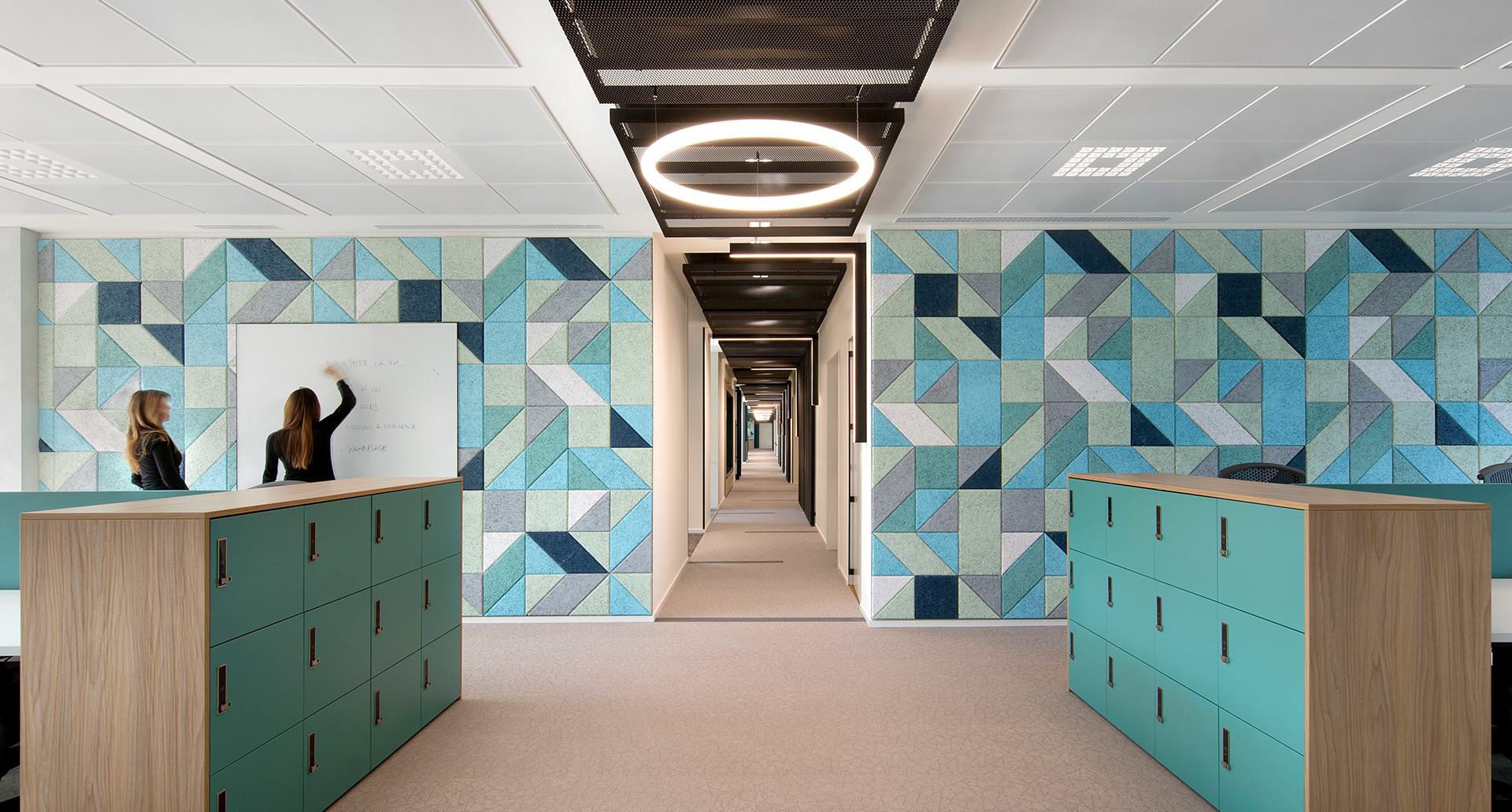 Modern office corridor with geometric teal-patterned walls, circular Artemide pendant light on ceiling, and reception desks with staff.
