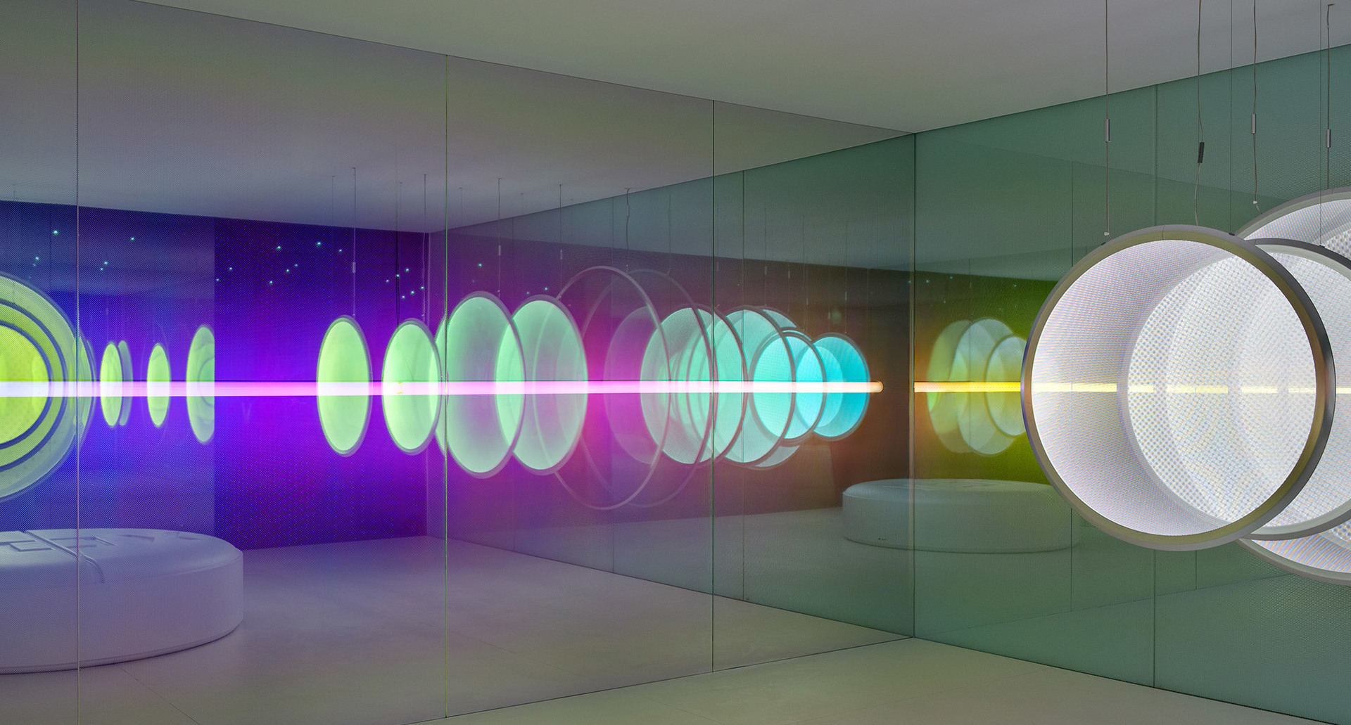 Sala specchiata immersiva con anelli luminosi al neon colorati in viola, verde e ciano che creano riflessi infiniti lungo le pareti