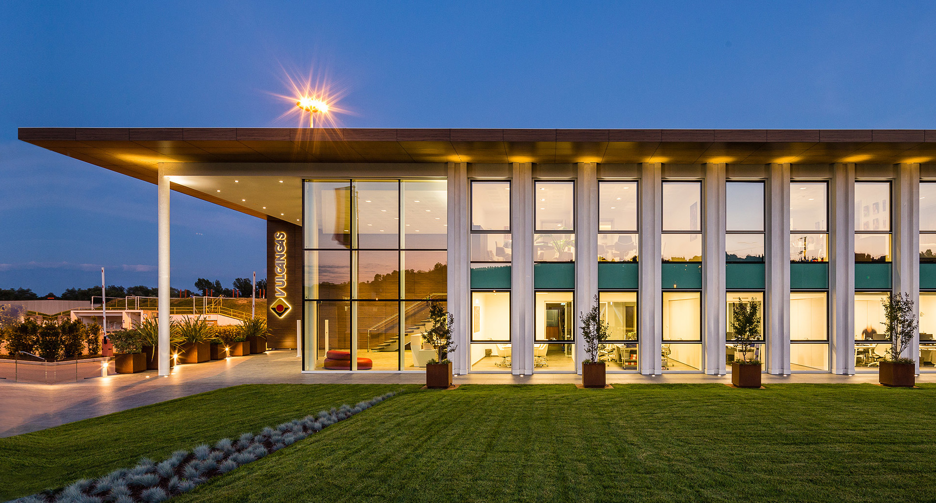 Edificio moderno con facciata in vetro illuminato al crepuscolo, con calda luce interna e faretti a soffitto su un tetto piano esteso.