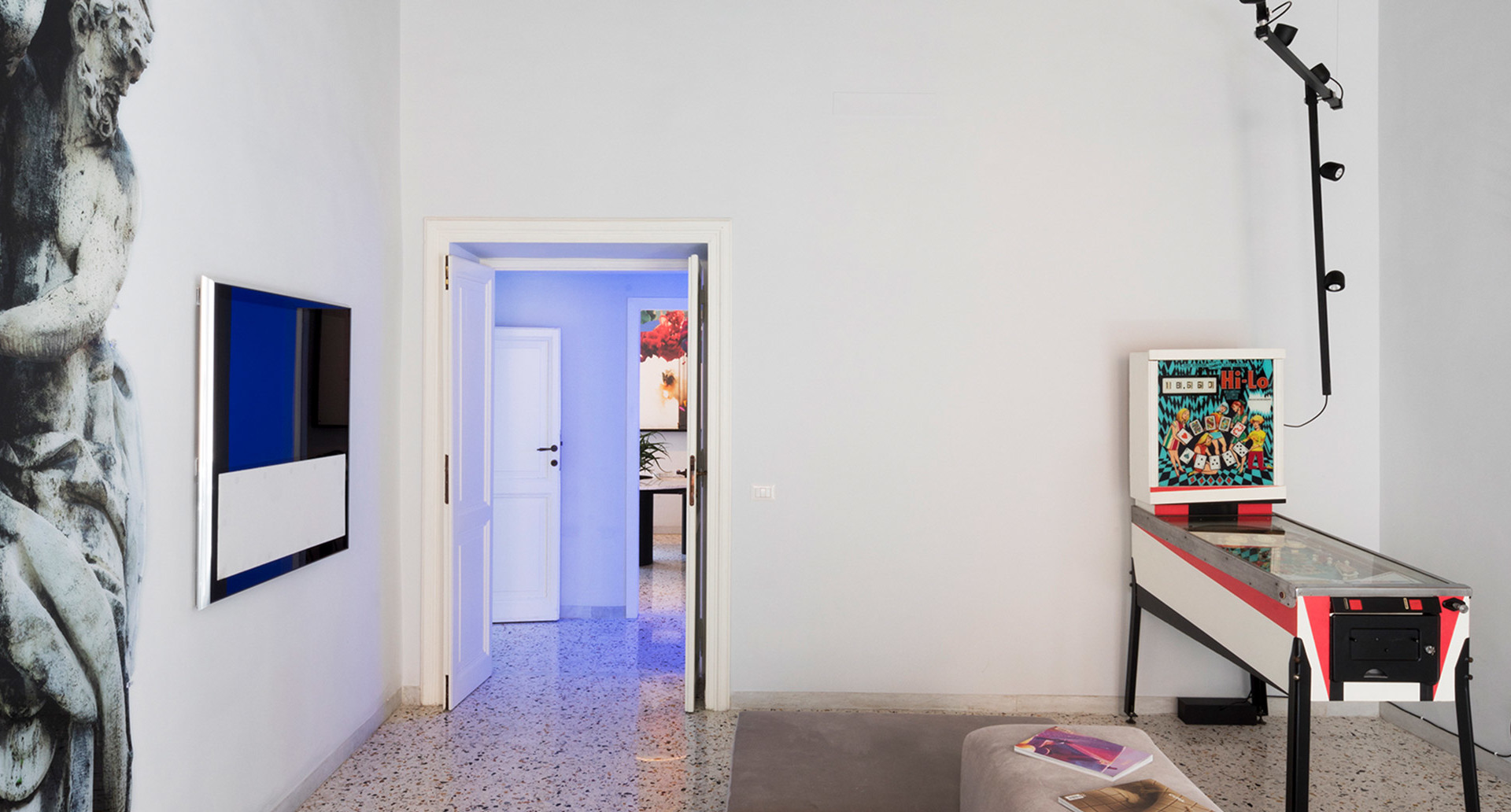 Stanza bianca moderna con flipper, opere d'arte a parete, TV e lampada da terra nera con faretti regolabili multipli su palo verticale