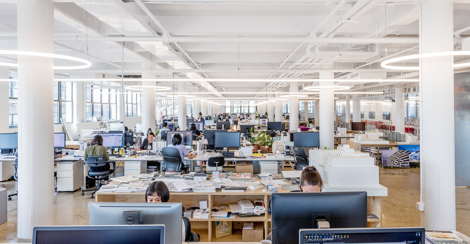 Open-plan office with employees at workstations, illuminated by linear pendant and ceiling-mounted fluorescent light fixtures.