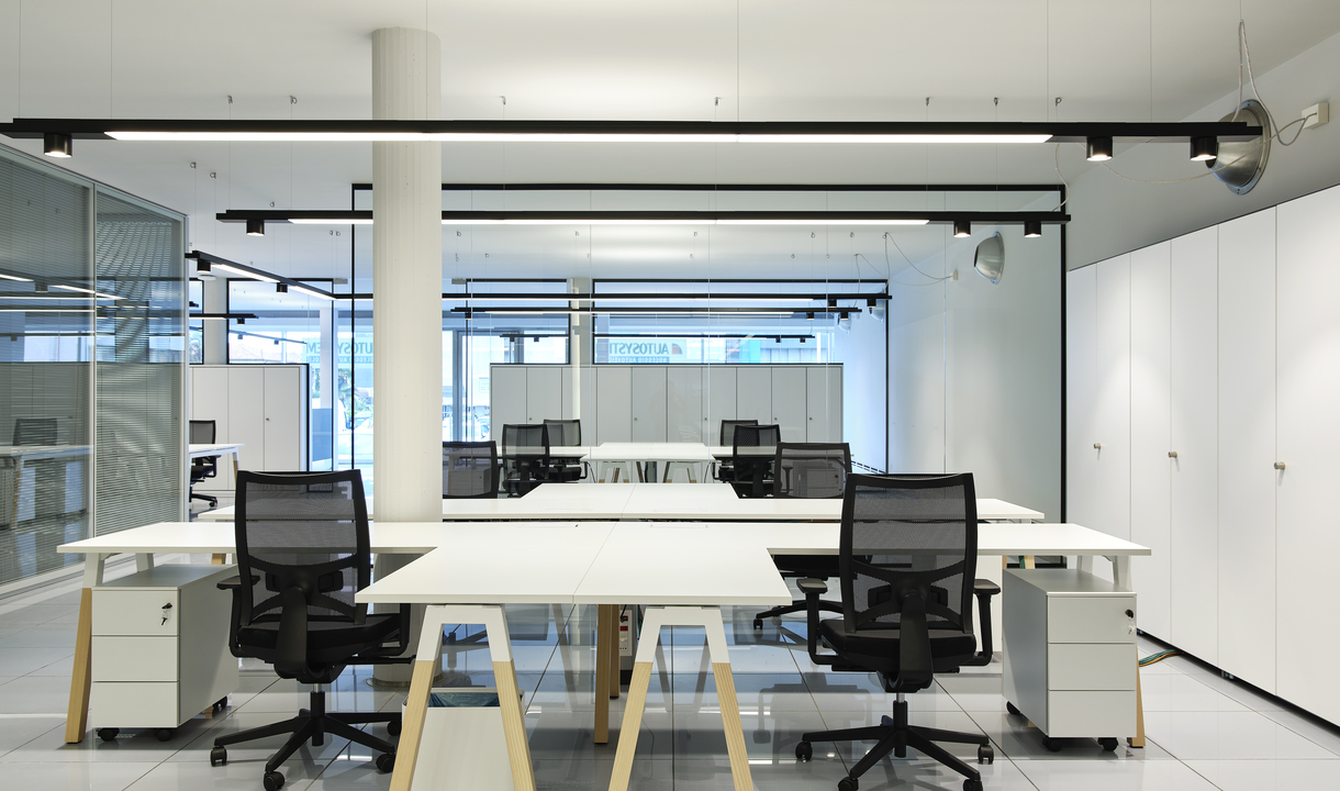 Ufficio open space moderno con postazioni bianche, sedie a rete e sistema di illuminazione a binario nero Artemide montato sul soffitto.