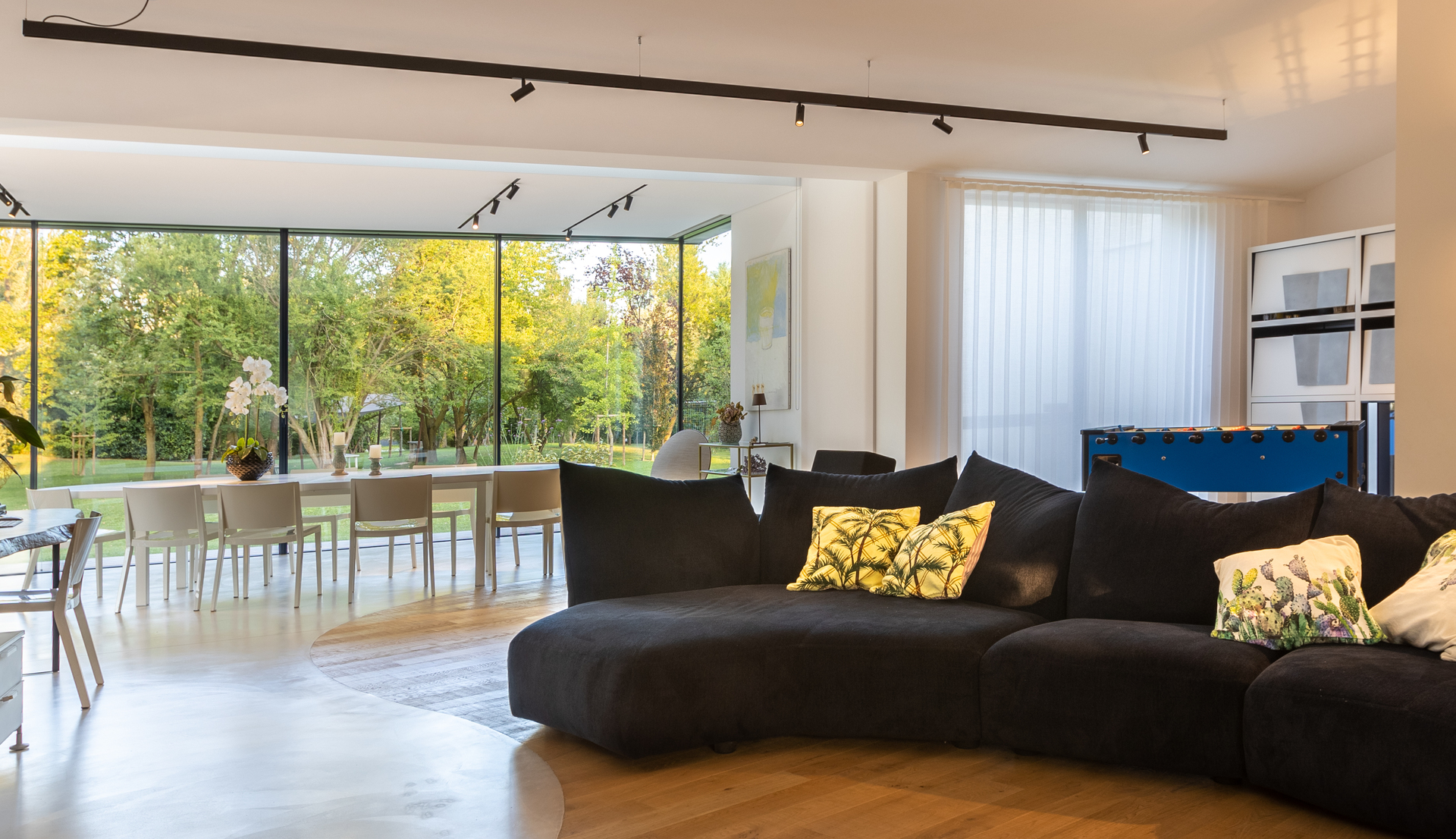 Soggiorno moderno open space con faretti su binario a soffitto, ampio divano curvo nero, finestre a tutta altezza con vista su un giardino rigoglioso