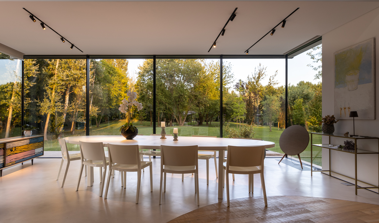 Sala da pranzo moderna con tavolo ovale bianco, sedie beige e faretti su binario nero a soffitto, affacciata su un giardino lussureggiante attraverso vetrate a tutta altezza.