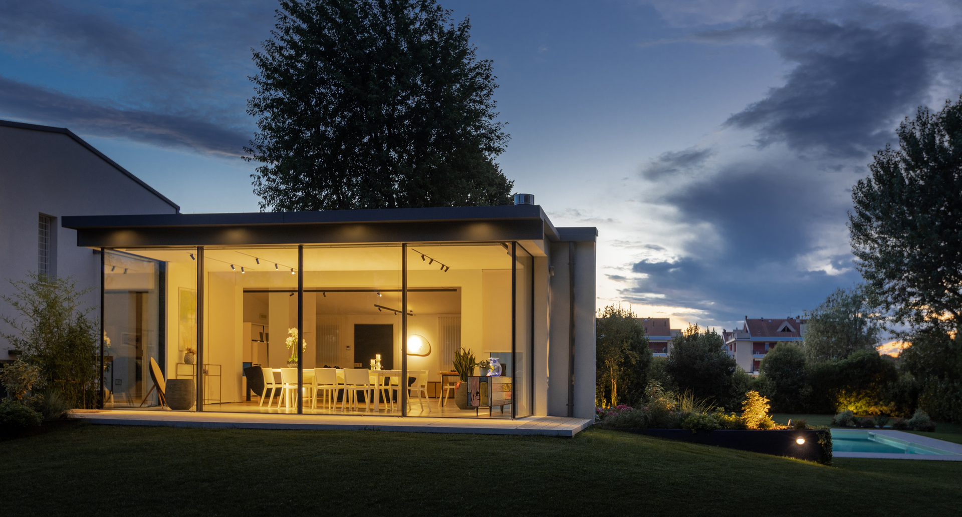 Casa moderna con pareti in vetro al crepuscolo, interno caldo con faretti su binario e lampada a sospensione visibili attraverso finestre a tutta altezza