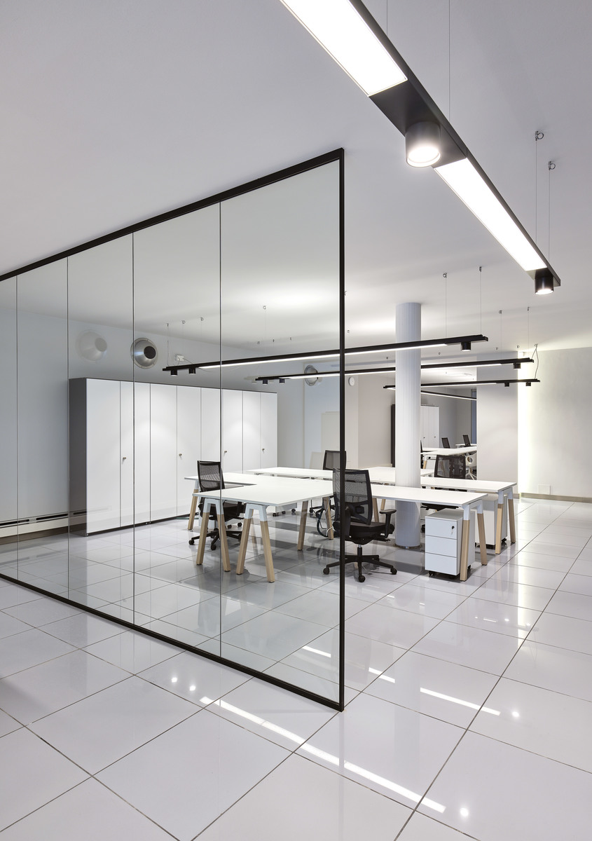 Ufficio open space moderno con pareti divisorie in vetro, postazioni bianche e lampade lineari LED Artemide a soffitto per illuminazione uniforme