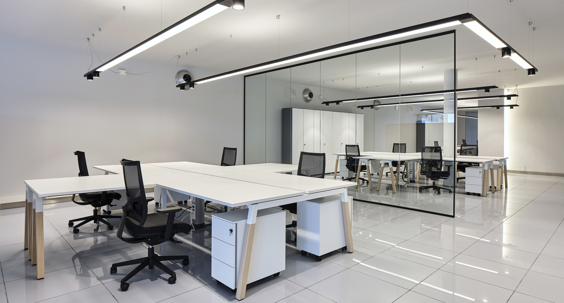 Ufficio open space moderno con postazioni bianche, sedie a rete e lampade lineari LED Artemide a soffitto che illuminano sala riunioni con pareti in vetro.