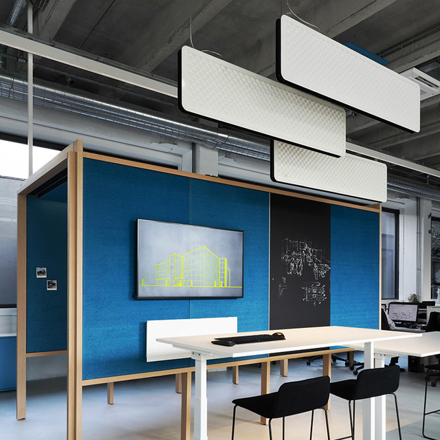 Pannelli acustici a sospensione Artemide con illuminazione integrata sopra una postazione ufficio moderna con pareti divisorie blu e monitor.