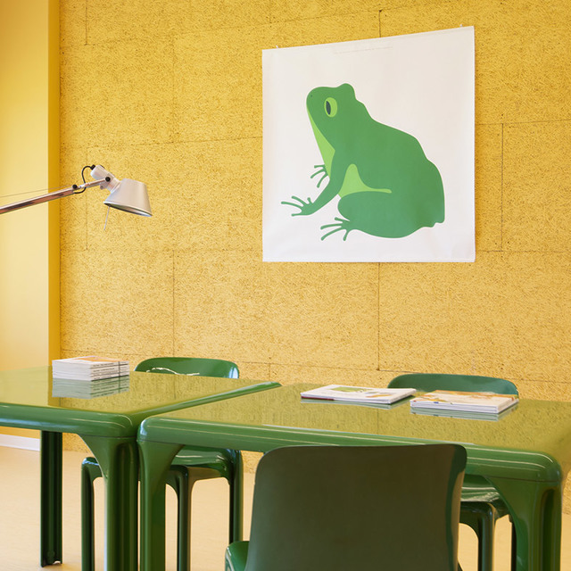 Lampada da tavolo Artemide Tolomeo su scrivania verde con sedie abbinate, parete in sughero giallo e opera d'arte con rana verde sullo sfondo