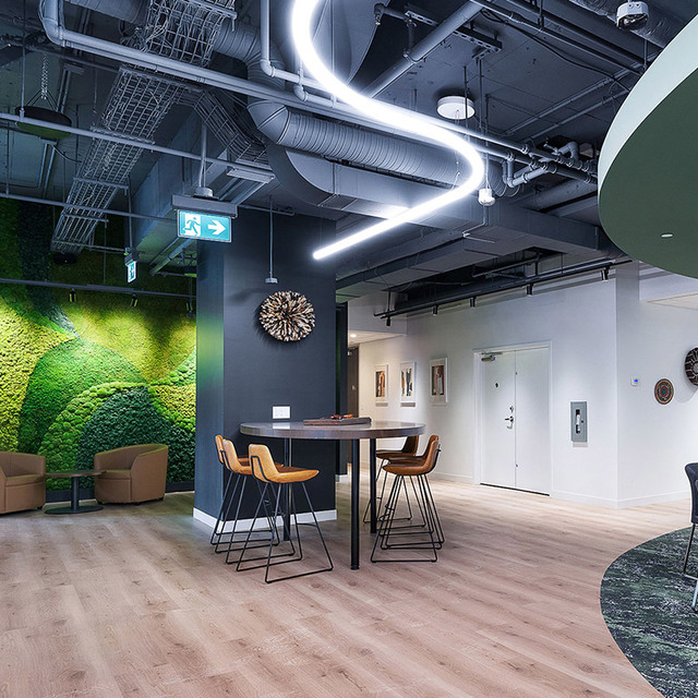 Lounge ufficio moderno con lampada a sospensione LED lineare sinuosa, parete di muschio verde, condotti a vista, tavolo alto e pavimento in legno caldo