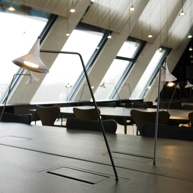 Lampade da terra Artemide con paralumi conici bianchi su strutture angolari nere illuminano un'area lounge moderna con grandi finestre inclinate.