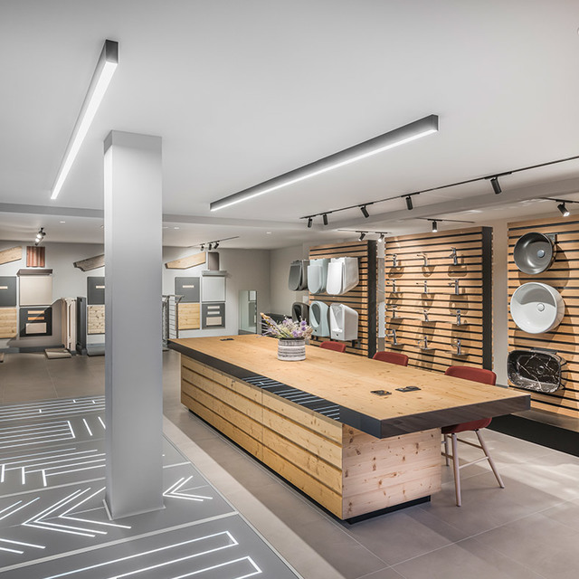 Interno di showroom con plafoniere lineari LED Artemide e faretti su binario che illuminano un banco centrale in legno e parete espositiva.