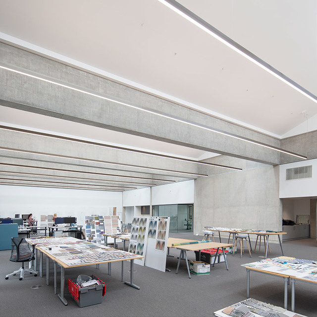 Studio open space con luci a soffitto LED lineari che illuminano tavoli da lavoro coperti di disegni progettuali e campioni di materiali.