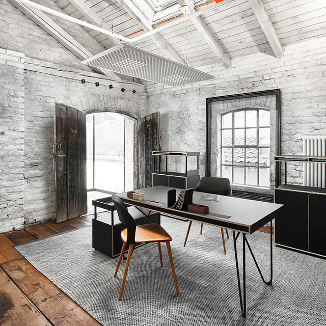 Ufficio in stile industriale con pareti in mattoni a vista, lucernario, illuminazione a binario, scrivania nera con gambe a forcina e sedie moderne su tappeto grigio