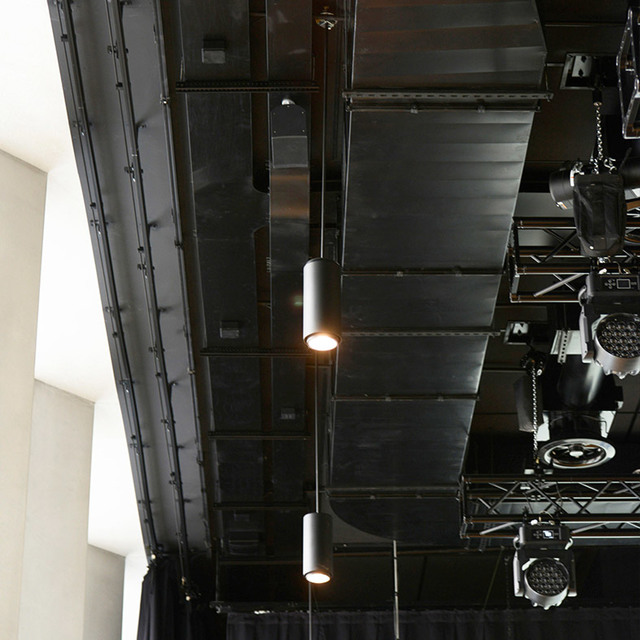 Due lampade a sospensione cilindriche nere appese a un soffitto industriale scuro con tralicci metallici e attrezzatura per illuminazione scenica.