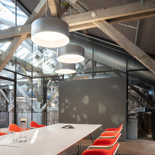 Tre lampade a sospensione cilindriche Artemide grigie illuminano un tavolo conferenze bianco con sedie arancioni in un loft industriale.