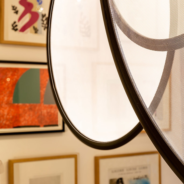 Primo piano di una lampada circolare con struttura in metallo nero e diffusore a rete, che riflette opere incorniciate su una parete galleria