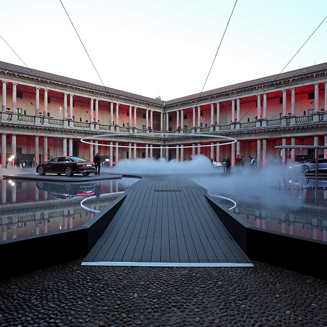 Installazione luminosa ad anello circolare Artemide su una passerella nebbiosa in un cortile neoclassico illuminato di rosso al crepuscolo