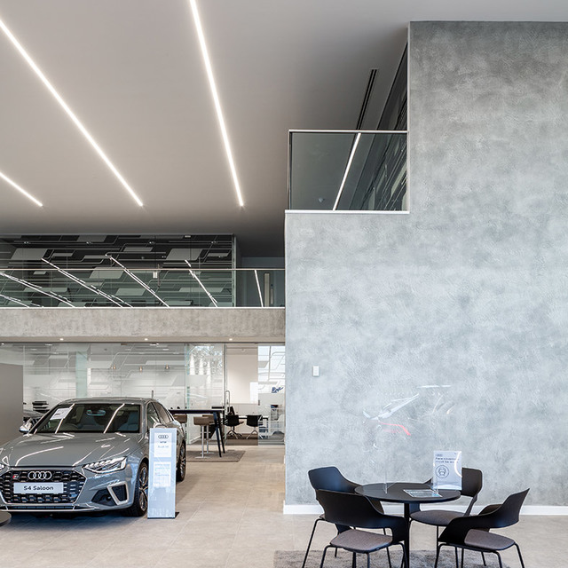 Showroom Audi con luci LED lineari a incasso nel soffitto che illuminano una A4 Saloon grigia in esposizione e un'area salotto.