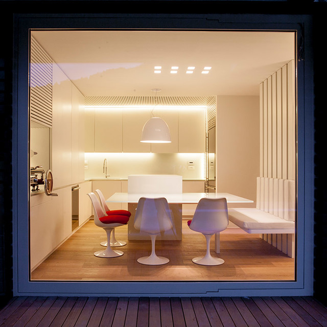 Modern dining room viewed through large window, featuring a white pendant lamp over a table with tulip chairs and warm LED strip lighting.