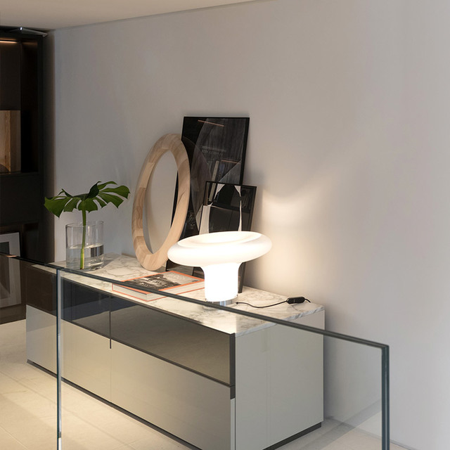 Lampada da tavolo Artemide in vetro bianco a forma di fungo illuminata su una credenza con piano in marmo, abbinata a uno specchio rotondo e una pianta monstera