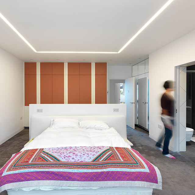 Camera da letto moderna con luci lineari LED da incasso Artemide a soffitto che formano un motivo geometrico, illuminando un letto con copriletto colorato.